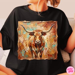 May include: Black t-shirt featuring a square, textured image of a longhorn steer. The steer is brown with long horns, set against a background of brown, teal, and gold paint strokes. The shirt is a casual style.