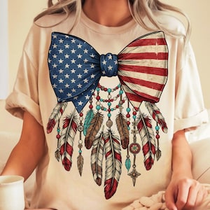Peut inclure: T-shirt crème avec un grand motif de nœud papillon représentant le drapeau américain. En dessous, des plumes décoratives rouges, blanches, bleues et marron, avec des perles.