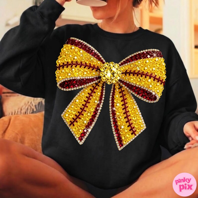 May include: Black sweatshirt with a large, glittering bow design. The bow is adorned with yellow and red rhinestones, and a central jewel. The bow's outline is also accented with clear rhinestones. The sweatshirt is a casual garment.