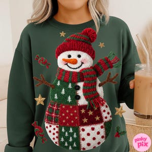 May include: A dark green sweatshirt featuring a whimsical snowman design. The snowman has a red knit hat and scarf, a carrot nose, and a patchwork body with green, red, and white squares. Gold star and candy accents surround the snowman.