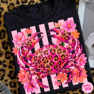 May include: Black crew neck t-shirt featuring a vibrant crab design. The crab has a leopard print pattern on its shell and claws, surrounded by pink and orange flowers. The background has pink stripes.