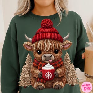 May include: Green sweatshirt featuring a cartoon Highland cow wearing a red knit hat and scarf. The cow is holding a red mug with a snowflake design and whipped cream. Two small Christmas trees flank the cow. The sweatshirt has a crew neck.