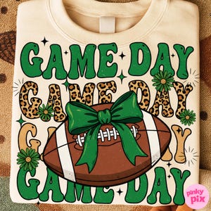 May include: Cream-colored sweatshirt with a football graphic and the words "GAME DAY" in green, leopard print, and tan. A green bow is tied around the football. The design includes green flowers and stars.