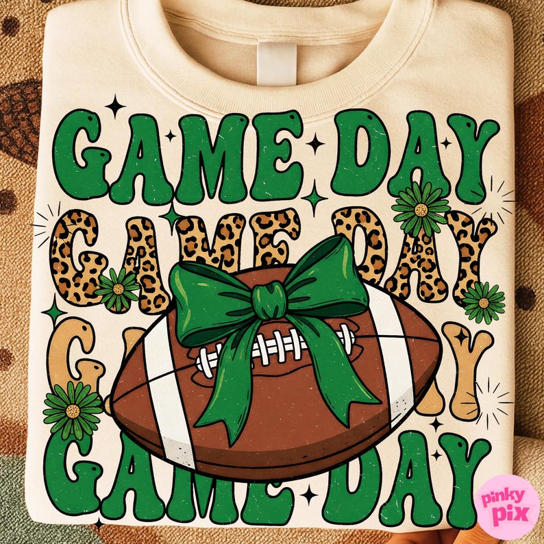 May include: Cream-colored sweatshirt with a football graphic and the words "GAME DAY" in green, leopard print, and tan. A green bow is tied around the football. The design includes green flowers and stars.