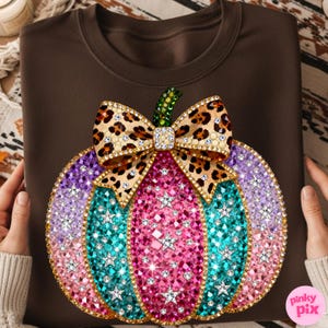 Op de afbeelding: Een donkerbruin sweatshirt met een grote pompoenafbeelding bestaande uit veelkleurige strass-steentjes in roze, taling en paarse tinten. De pompoen is versierd met een strik met luipaardprint en een groene steel, bezet met fonkelende sterren.