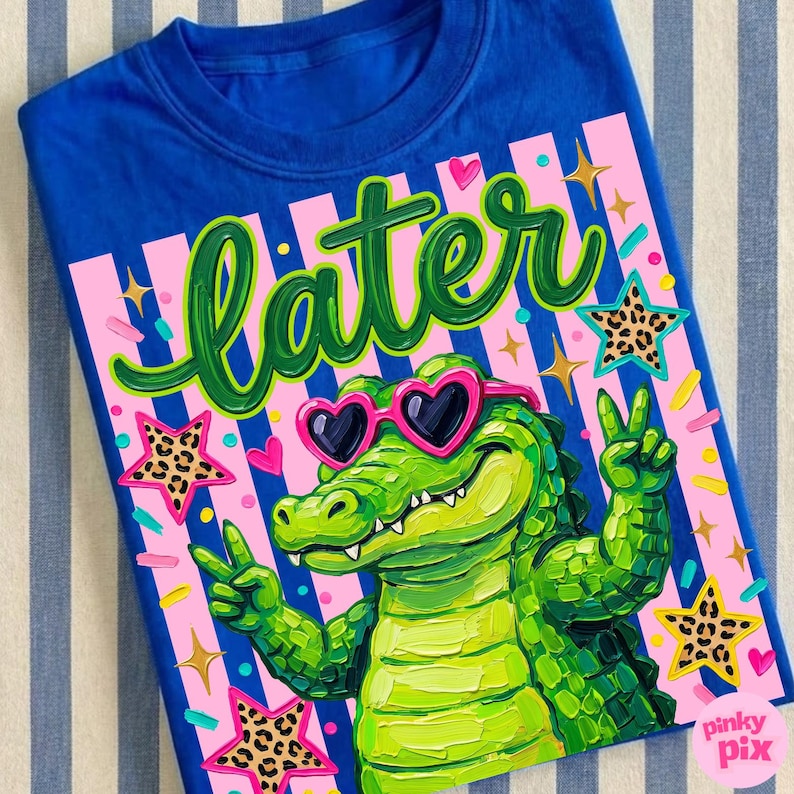 May include: Blue t-shirt with a graphic of a green alligator wearing heart-shaped sunglasses and giving a peace sign. The word "later" is written in green script above the alligator. Pink and gold accents complete the design.
