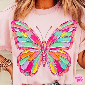 May include: A light pink t-shirt featuring a large, colorful butterfly design. The butterfly has a textured, painted appearance with shades of pink, green, orange, yellow, and purple. The shirt is a crew neck style.