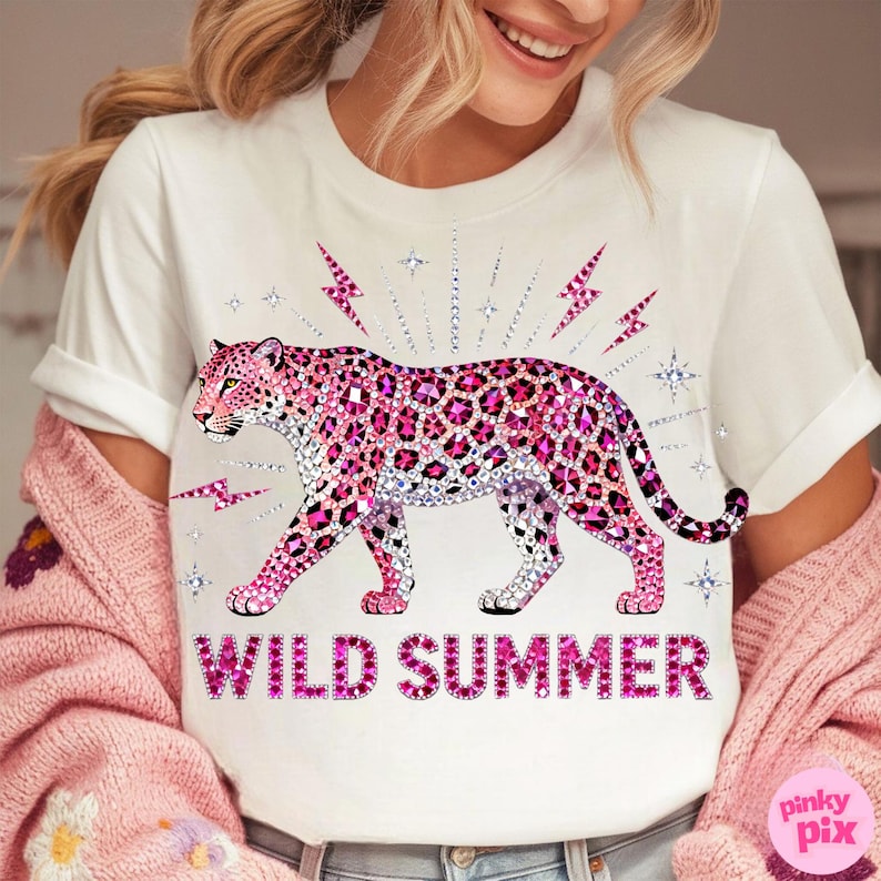 May include: White t-shirt featuring a pink leopard design with a rhinestone pattern. The leopard is surrounded by pink lightning bolts and stars. The words "WILD SUMMER" are printed in pink below the leopard.