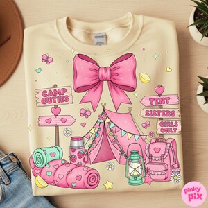 May include: Cream-coloured sweatshirt with a pink bow and camping-themed graphics. Features a pink tent, sleeping bag, backpack, and signs that read "Camp Cuties," "Tent Sisters," and "Girls Only." Includes butterflies, hearts, and stars.