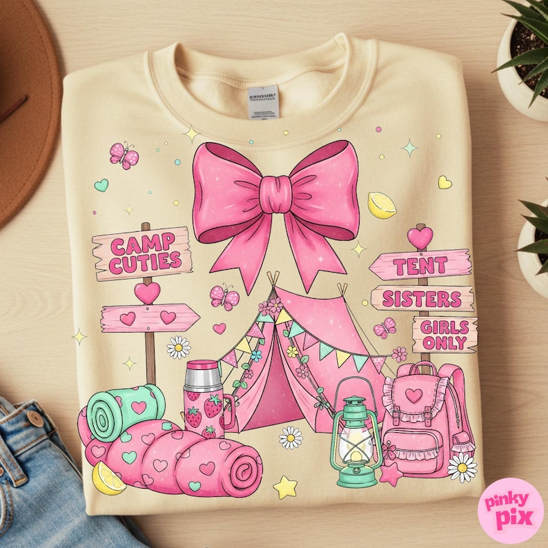 May include: Cream-coloured sweatshirt with a pink bow and camping-themed graphics. Features a pink tent, sleeping bag, backpack, and signs that read "Camp Cuties," "Tent Sisters," and "Girls Only." Includes butterflies, hearts, and stars.