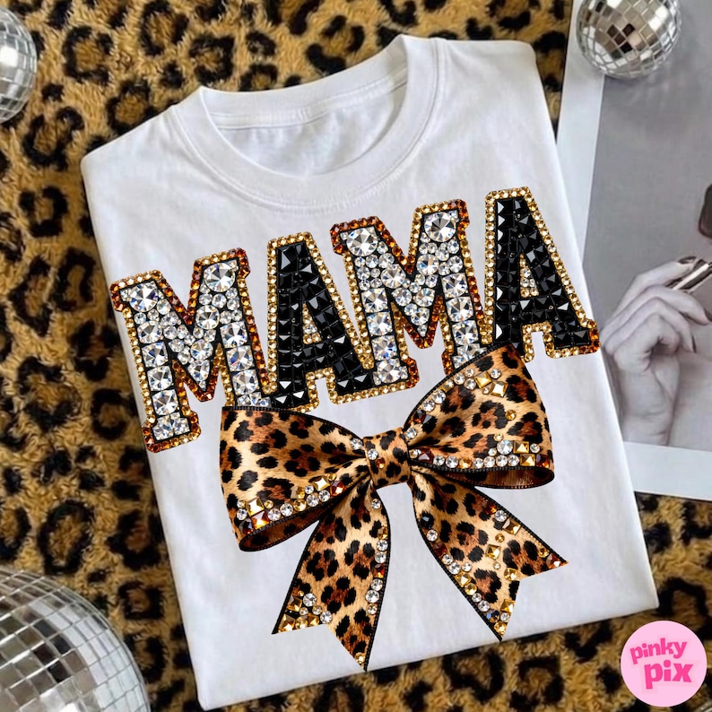 May include: White t-shirt with the word "MAMA" in rhinestone-studded letters, accented with black gems. A leopard print bow with rhinestone detailing is centered below the text. The shirt is displayed on a leopard print surface.