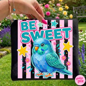 May include: Black tote bag featuring a colorful illustration of a blue parakeet with the words "BE SWEET" in teal and white. The design includes pink and black stripes, stars, and flowers. The bag is held against a background of green grass and flowers.