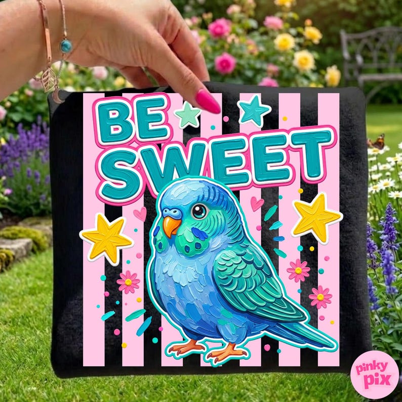 May include: Black tote bag featuring a colorful illustration of a blue parakeet with the words "BE SWEET" in teal and white. The design includes pink and black stripes, stars, and flowers. The bag is held against a background of green grass and flowers.
