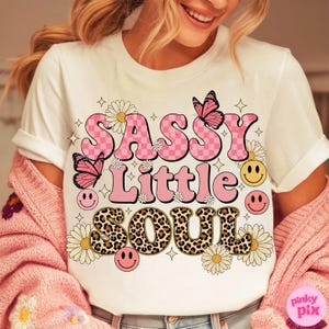 May include: White t-shirt with the text "SASSY LITTLE SOUL" in pink, leopard print, and checkerboard patterns. The design includes butterflies, daisies, smiley faces, and stars. The shirt is part of a clothing collection.
