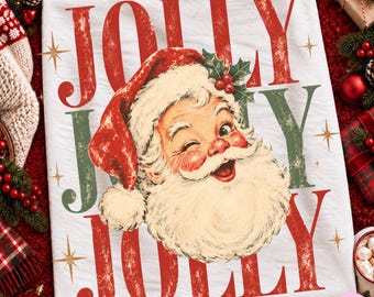 May include: A white t-shirt featuring a vintage-style illustration of a winking Santa Claus wearing a red hat with holly berries. The text JOLLY is printed in large, distressed red and green letters above and below the Santa graphic.
