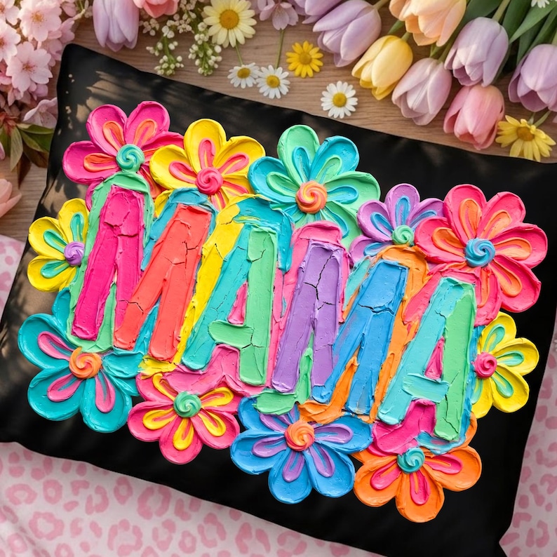 May include: Black decorative pillow with the word "MAMA" in colorful, textured letters, surrounded by vibrant, painted flowers in shades of pink, yellow, blue, and orange. The pillow is a decorative home accent.