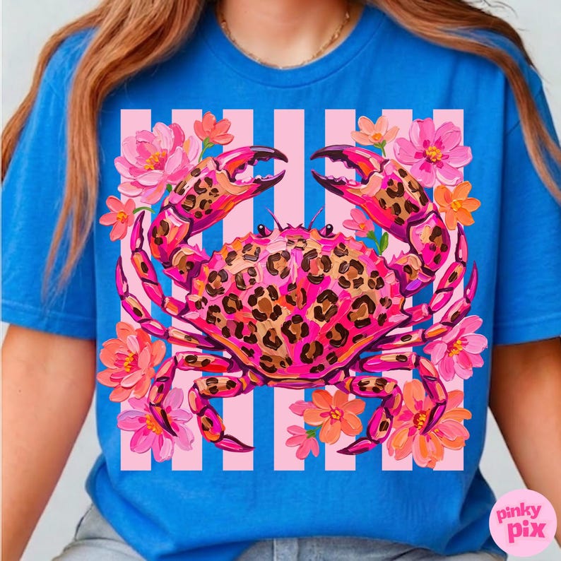 May include: A blue t-shirt featuring a vibrant crab design. The crab is pink with a leopard print pattern, surrounded by pink and orange flowers. The background has vertical pink and white stripes.
