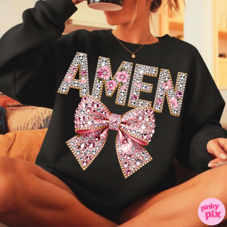 May include: Black sweatshirt featuring the word "AMEN" in gold and rhinestone lettering, with pink floral details. A large pink rhinestone bow is centered below the word. The sweatshirt has a crew neck.