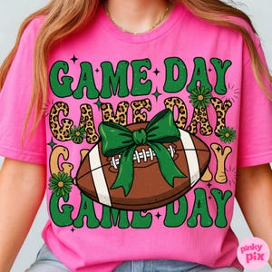 May include: Pink t-shirt with the words "GAME DAY" in green, leopard print, and gold lettering. A brown football with a green bow is centered on the shirt, with green flowers and stars completing the design.