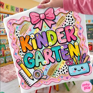 May include: A colorful kindergarten sign with the word "KINDERGARTEN" in large, multi-colored letters. The sign features a pink bow, a pencil, and a pencil case with school supplies. The sign is decorated with stars, flowers, and gold glitter accents.