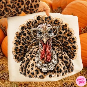 May include: A white sweatshirt featuring a graphic of a turkey with leopard print feathers, wearing leopard print cat-eye glasses and a large, clear crystal statement necklace. The turkey has a red wattle and a detailed, textured appearance.