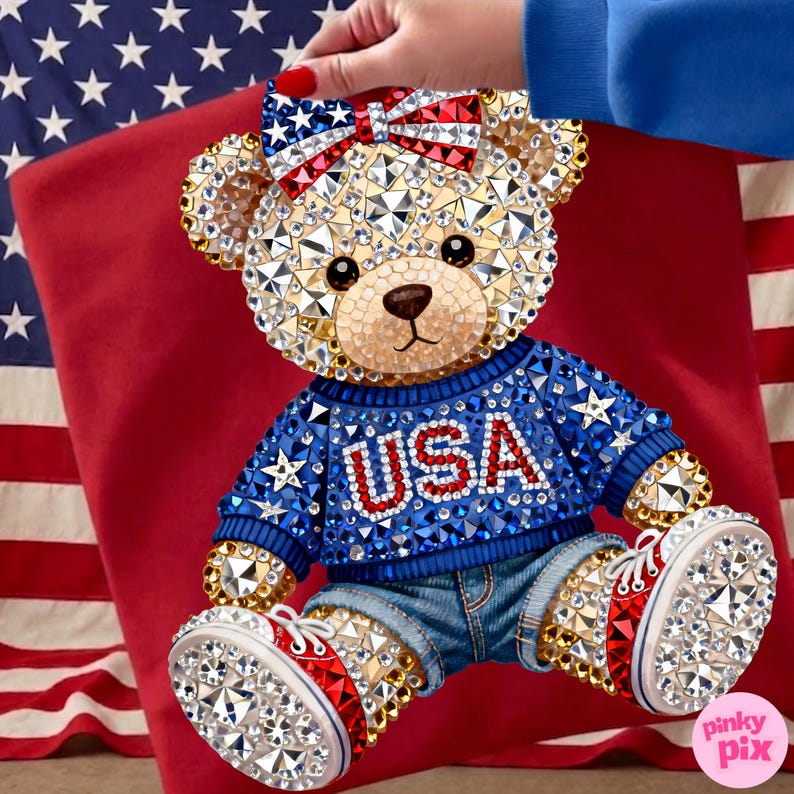 May include: A bedazzled teddy bear wearing a blue sweater with "USA" in red letters, denim shorts, and red, white, and blue sneakers. The bear has a bow with a star pattern. The background includes a red pillow and an American flag.