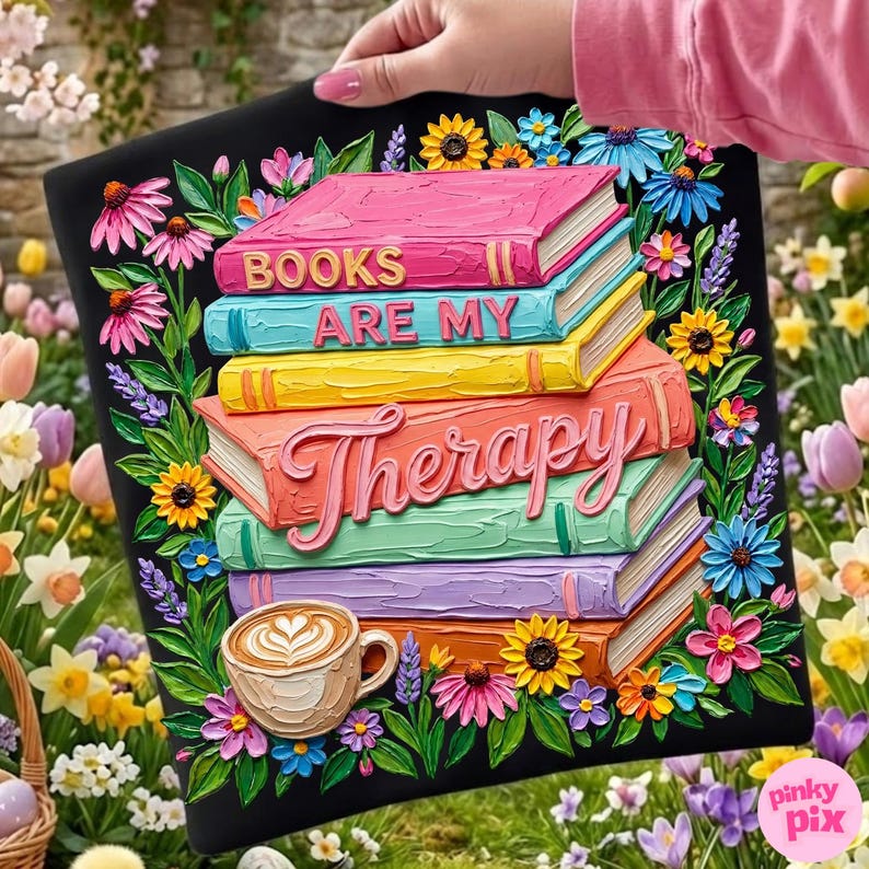 May include: Black tote bag featuring a stack of colorful books with the words "BOOKS ARE MY Therapy". The design includes a coffee cup and floral accents in pink, yellow, blue, and green. The bag is held against a background of flowers.