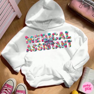 May include: A white hooded sweatshirt with the words "MEDICAL ASSISTANT" in colorful floral letters. A light blue bow is between the words. The sweatshirt is on a wooden surface with pink sneakers and a book.