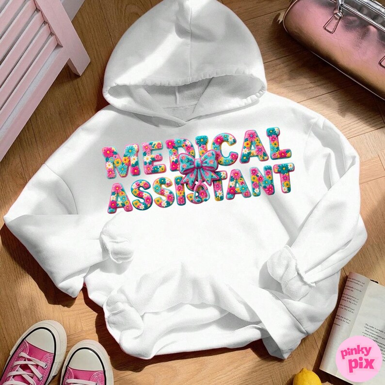 May include: A white hooded sweatshirt with the words "MEDICAL ASSISTANT" in colorful floral letters. A light blue bow is between the words. The sweatshirt is on a wooden surface with pink sneakers and a book.