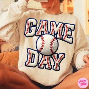 May include: Cream-colored sweatshirt with the words "GAME DAY" in white, outlined in navy blue with red stitching, and a baseball graphic. The baseball is white with red stitching. The sweatshirt is a casual, sporty style.