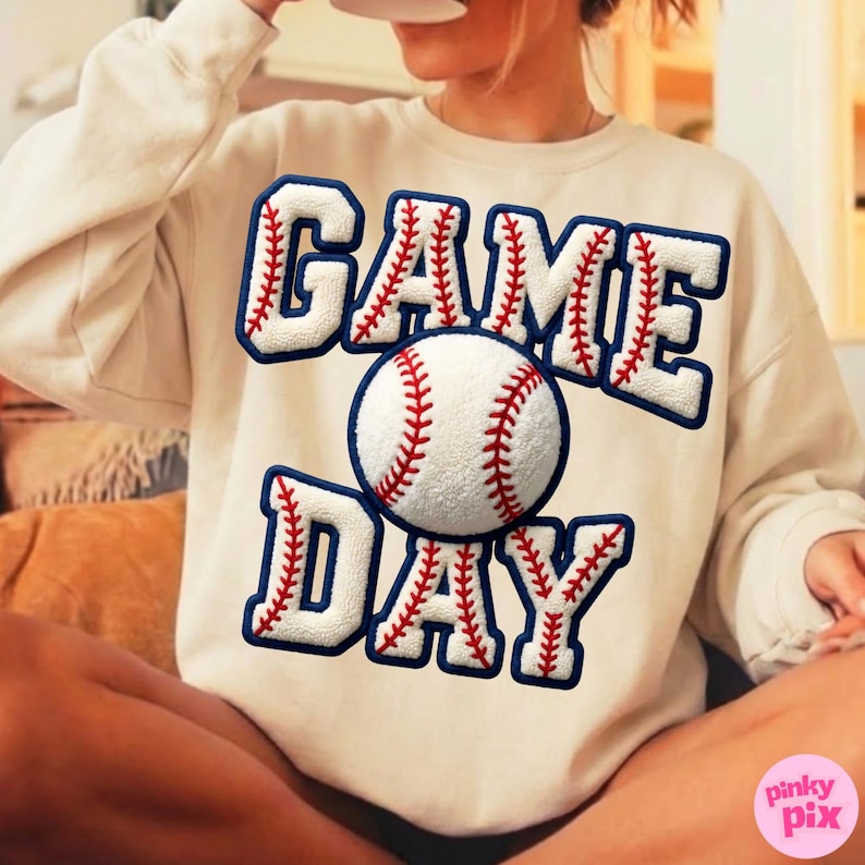 May include: Cream-colored sweatshirt with the words "GAME DAY" in white, outlined in navy blue with red stitching, and a baseball graphic. The baseball is white with red stitching. The sweatshirt is a casual, sporty style.