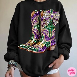 May include: Black sweatshirt featuring a vibrant graphic of jeweled cowboy boots. The boots are adorned with purple, green, and gold mosaic-style embellishments, and a large, sparkling bow. The design is detailed with rhinestones.