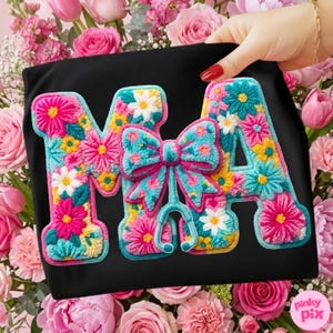 May include: Black t-shirt with the embroidered letters "MA" in a floral design. The letters are filled with pink, yellow, and white flowers, and a turquoise bow and stethoscope are in the center.