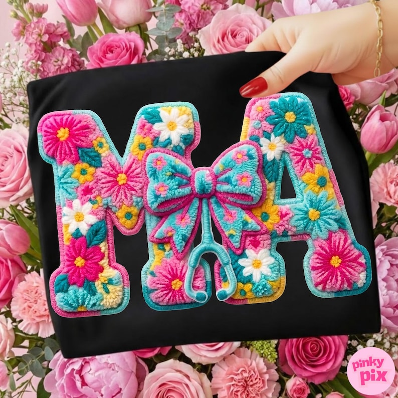 May include: Black t-shirt with the embroidered letters "MA" in a floral design. The letters are filled with pink, yellow, and white flowers, and a turquoise bow and stethoscope are in the center.