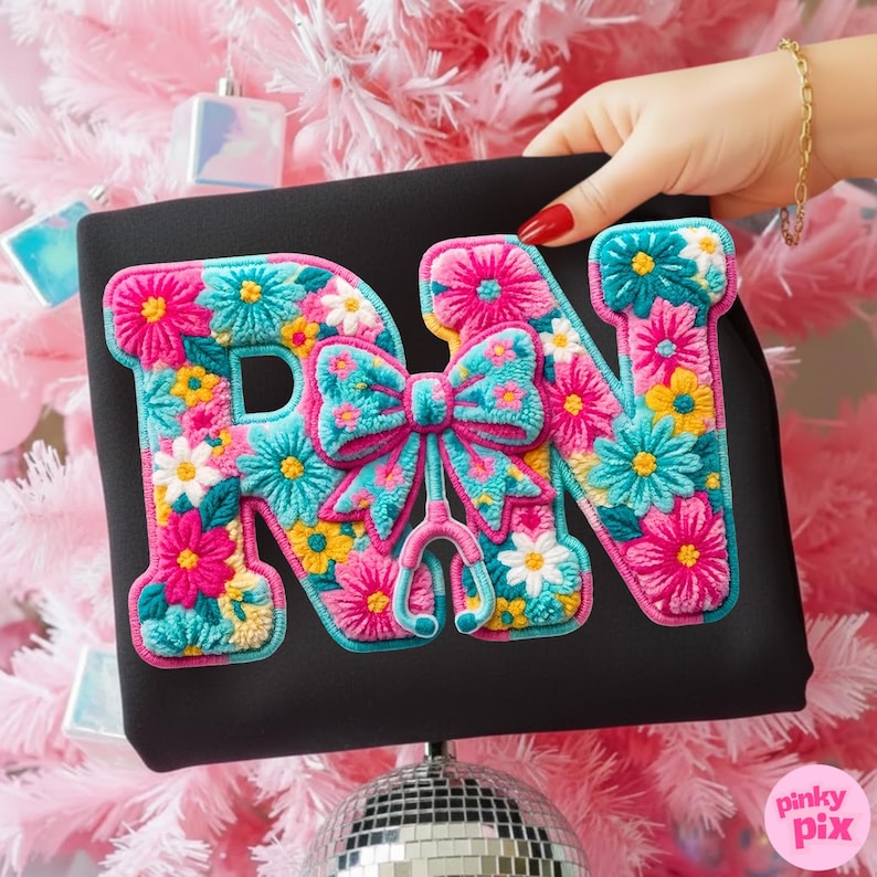 May include: Black sweatshirt featuring a colorful floral design spelling out "RN" with a bow and stethoscope. The letters are filled with pink, blue, yellow, and white flowers. The sweatshirt is held up against a pink Christmas tree.