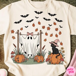 May include: Cream-colored t-shirt featuring a Halloween-themed design. The graphic includes a ghost with a pink bow, pumpkins, a black cat wearing a witch hat, bats, and autumn leaves. The design is whimsical and seasonal.