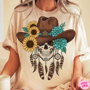 Op de afbeelding: Beige T-shirt met een gedetailleerde afbeelding. De afbeelding toont een schedel met een bruine cowboyhoed versierd met zonnebloemen, turquoise bloemen en een ster. Dromenvangers en veren hangen onder de schedel en cre&euml;ren een artistiek ontwerp.