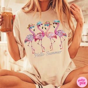 May include: Beige t-shirt featuring a rhinestone design of four pink flamingos wearing hats and sunglasses. The phrase "Hello Summer" is written below the flamingos in a sparkly font. The shirt is a casual, relaxed fit.