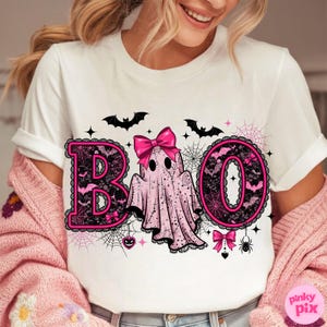 Puede incluir: Camiseta blanca con un dise&ntilde;o tem&aacute;tico de Halloween. La palabra "BOO" est&aacute; escrita con letras negras tipo encaje delineadas en rosa. Un fantasma rosa con un lazo est&aacute; en el centro, con murci&eacute;lagos y telara&ntilde;as.