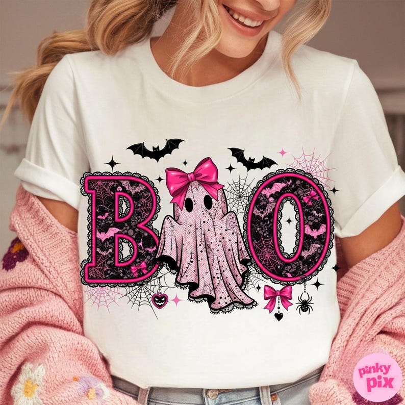 Puede incluir: Camiseta blanca con un dise&ntilde;o tem&aacute;tico de Halloween. La palabra "BOO" est&aacute; escrita con letras negras tipo encaje delineadas en rosa. Un fantasma rosa con un lazo est&aacute; en el centro, con murci&eacute;lagos y telara&ntilde;as.