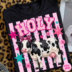 May include: Black t-shirt with a graphic of a cow wearing a leopard print bow and the word "HOLY" in pink letters. The design includes pink stars, flowers, and stripes.
