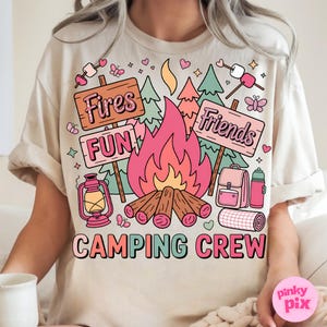 May include: Beige t-shirt with a colorful camping-themed graphic. The design includes a campfire, trees, signs that say "Fires", "Fun", and "Friends", and the words "Camping Crew". Other elements include a lantern, backpack, and marshmallows.