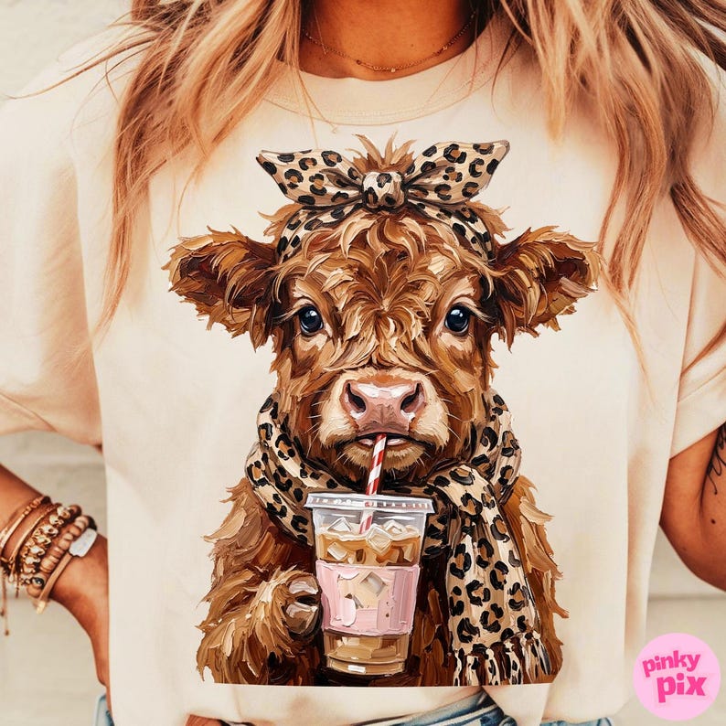 May include: A beige t-shirt featuring a brown cow with a leopard print bow, scarf, and a drink with a red and white striped straw. The cow has a pink nose and is drinking from a clear cup with ice and a pink sleeve.