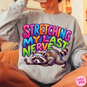 May include: A heather gray sweatshirt with the phrase "STRETCHING MY LAST NERVE" in colorful bubble letters. A cartoon raccoon wearing a headband is stretching below the text. The sweatshirt is a casual, comfortable style.