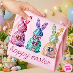 May include: A light pink t-shirt featuring three jeweled bunny designs in pink, blue, and green, each with a heart-shaped gem. The words "happy easter" are written in pink rhinestones. Easter-themed decor surrounds the shirt.