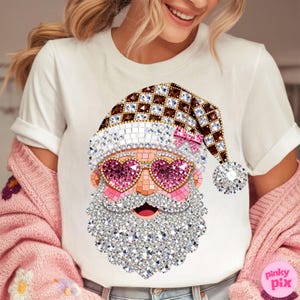 May include: A white short-sleeved t-shirt featuring a graphic of Santa Claus wearing heart-shaped pink glasses and a brown and white chequered hat. The design is rendered in a sparkling, faceted style with a pink bow on the hat and a white beard.