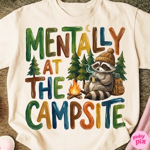 May include: Cream-colored t-shirt with the text "MENTALLY AT THE CAMPSITE" in colorful, block letters. A raccoon wearing a knit cap is depicted holding a mug near a campfire and pine trees. A backpack is behind the raccoon.