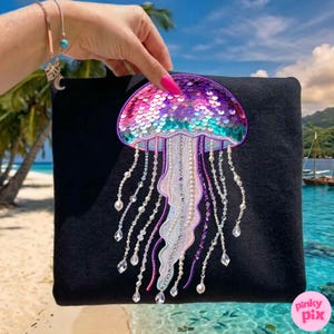 May include: Black clutch featuring a colorful sequined jellyfish design. The jellyfish has a pink, purple, and teal sequined cap, with beaded and crystal accents. The clutch is held against a tropical beach backdrop.