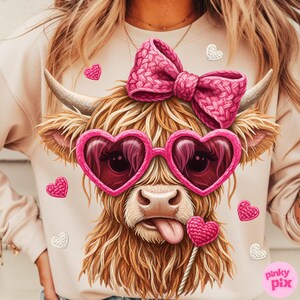 May include: A light beige sweatshirt featuring a whimsical illustration of a Highland cow. The cow has pink heart-shaped sunglasses, a pink bow, and a lollipop. Scattered around are pink and white heart-shaped embellishments. The text "pinky pix" is in the bottom right corner.