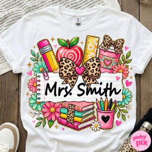 May include: White t-shirt featuring a colorful teacher-themed design. The design includes a pencil, apple, ruler, coffee cup, books, and pencils. The text "Mrs. Smith" is written on a lined paper. The design is surrounded by flowers and hearts.